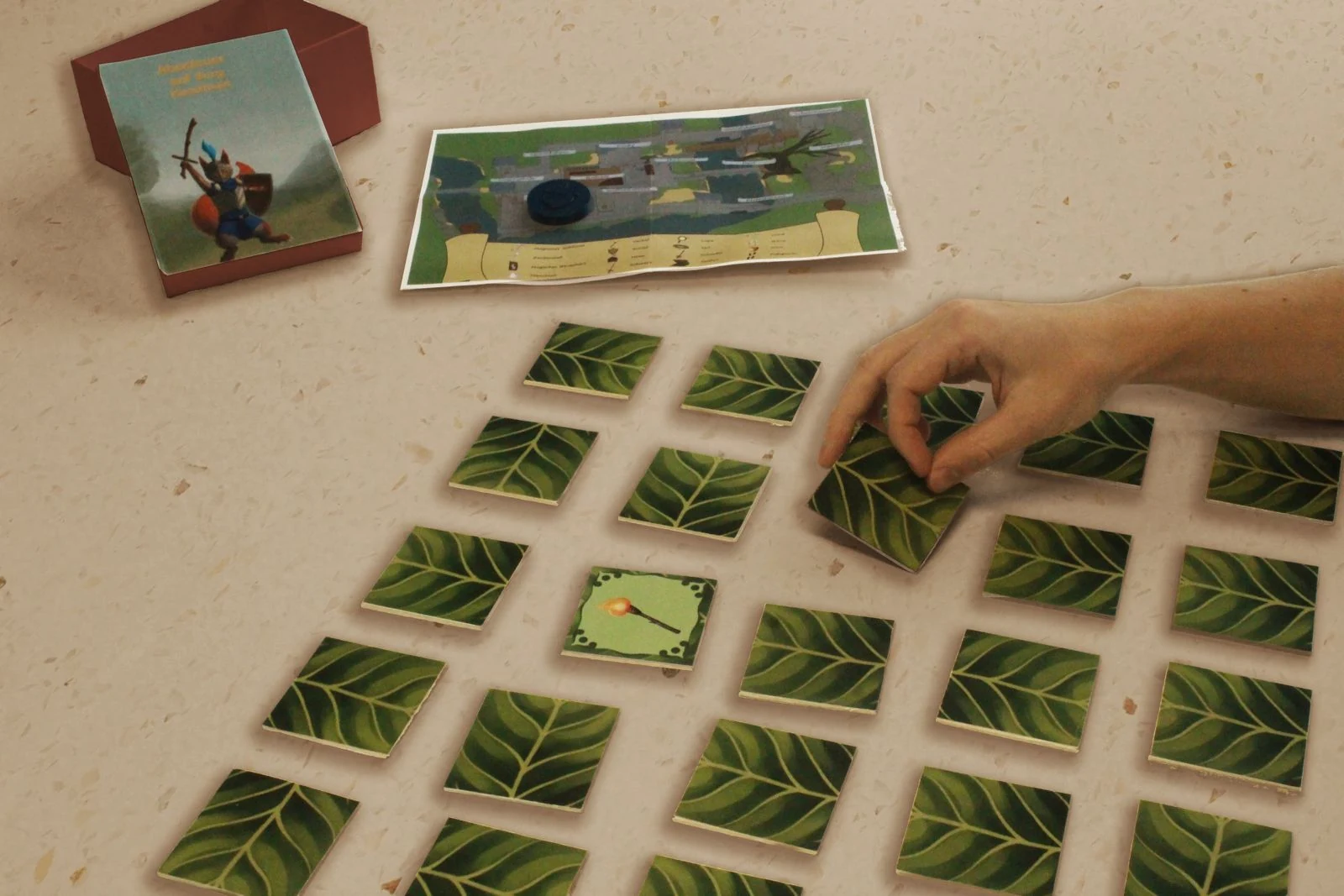 Ausgelegtes Memory-Spiel mit verdeckten Karten, Burgkarte, Schachtel und einer Hand beim Aufdecken